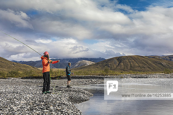 Caucasian woman in her 40's  wearing orange rain coat  casting her rod fly fishing for grayling in a pool of water off the Marsh Fork River  while a caucasian man in his 30's in the background wearing shorts is also fly fishing for grayling on a sunny summer day in the arctic  Brooks Range  Arctic National Wildlife Refuge; Alaska  United States of America