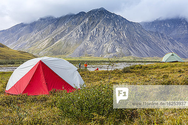 Red and grey backpacking tent in foreground  with another grey-green backpacking tent in middle ground  set up on tundra  with people (one caucasian man  two caucasian women) sitting and standing between the tents on a sunny day near the Marsh Fork river with mountains in the background rising into the clouds  Brooks Range  Arctic National Wildlife Refuge; Alaska  United States of America