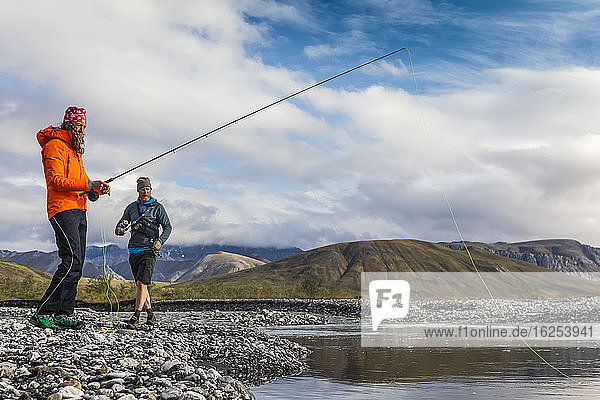 Caucasian woman in her 40's  wearing orange rain coat  casting her rod fly fishing for grayling in a pool of water off the Marsh Fork River  while a caucasian man in his 30's in the background wearing shorts is also fly fishing for grayling on a sunny summer day in the arctic  Brooks Range  Arctic National Wildlife Refuge; Alaska  United States of America