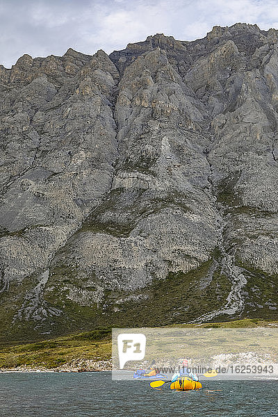 Zwei Bootsfahrer (ein kaukasischer Mann und eine kaukasische Frau) treiben an einem sonnigen Tag im Arctic National Wildlife Refuge in ihren gelben und blauen Packschiffen den Marsh Fork-Fluss hinunter  im Schatten der Brooks Range-Berge; Alaska  Vereinigte Staaten von Amerika