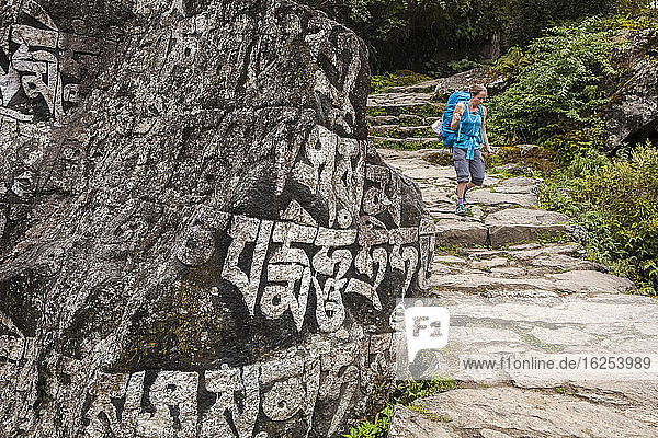 Eine Frau mit Rucksack geht einen Steinpfad entlang  streicht über eine Felswand mit Schriftzug; Nepal