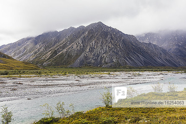 Launischer Tag in der Arktis  mit Wolken und Nebel in den Bergen  der Marsh Fork Fluss  der durch den Mittelgrund fließt  und Tundra im Vordergrund  an einem Sommertag in der Brooks Range  Arctic National Wildlife Refuge; Alaska  Vereinigte Staaten von Amerika