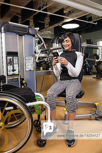 A paraplegic woman resting and texting on her smart phone after working out using an overhead press in a fitness facility; Sherwood Park  Alberta  Canada