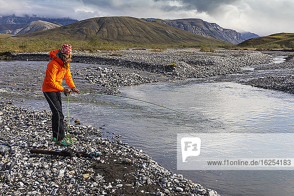 Caucasian woman in her 40's  wearing orange rain coat  fly fishing for grayling in a pool of water off the Marsh Fork river  on a sunny  summer day in the arctic  Brooks Range  Arctic National Wildlife Refuge; Alaska  United States of America