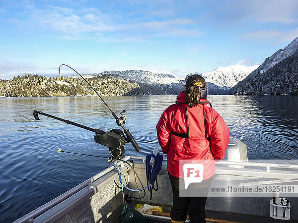 Rückansicht einer Frau  die im Boot neben einem Ausleger mit Angelruten steht und auf die Berge bei Homer schaut; Alaska  Vereinigte Staaten von Amerika