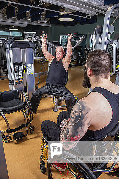 Ein querschnittsgelähmter Mann trainiert mit einer Überkopfpresse in einer Fitnesseinrichtung  während sein behinderter Freund zusieht; Sherwood Park  Alberta  Kanada