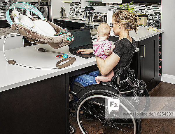Eine junge Mutter mit einer Rückenmarkverletzung kümmert sich um ihr Neugeborenes  während sie in der Küche an ihrem Computer arbeitet; Edmonton  Alberta  Kanada