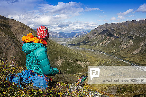 Wanderin in grüner  bauschiger Jacke  sitzt und genießt an einem sonnigen Sommertag den Blick aus der Vogelperspektive auf den Marsh Fork-Fluss und das Tal  Brooks Range  Arctic National Wildlife Refuge (ANWR); Alaska  Vereinigte Staaten von Amerika
