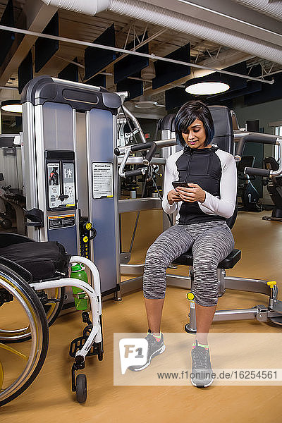 Eine querschnittsgelähmte Frau  die sich nach dem Training mit einer Überkopfpresse in einer Fitnesseinrichtung ausruht und mit ihrem Smartphone SMS schreibt; Sherwood Park  Alberta  Kanada