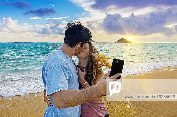 Ein Paar steht küssend am Strand von Lanakai auf der hawaiianischen Insel Oahu  während es ein Smartphone in der Hand hält und bei Sonnenuntergang ein Selbstporträt aufnimmt; Lanakai  Oahu  Hawaii  Vereinigte Staaten von Amerika