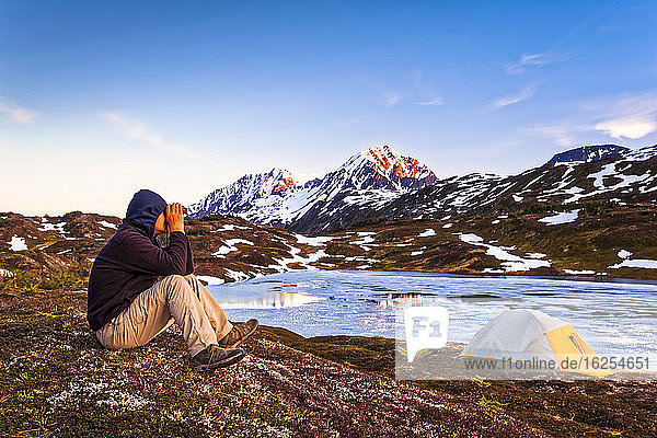 Ein Mann  der bei Sonnenuntergang mit einem Fernglas aus dem Lager am Lost Lake schaut. Gipfel der Auferstehung im Hintergrund. Chugach National Forest  Kenai-Halbinsel  Süd-Zentral-Alaska im Frühling; Seward  Alaska  Vereinigte Staaten von Amerika