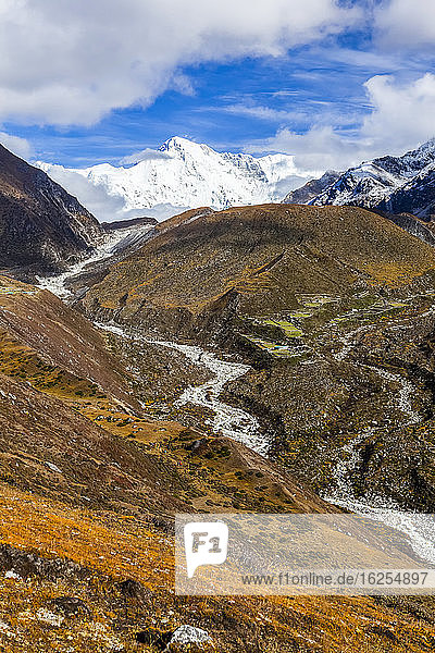 Clouds lifting  revealing a snow coated Cho Oyo rise above the Dudh Khosi river along the Gokyo trek on a sunny autumn day in the Himalayas  Sagarmatha National Park  Nepal. Trekkers are dwarfed by the landscape hiking up trail  and a teahouse sits among rock walled terraces on the other side of the river; Nepal