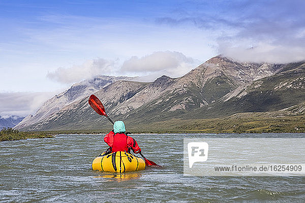 Woman wearing red drysuit and pfd  paddling yellow packraft boat down the Canning river through the Brooks Range  Arctic National Wildlife Refuge; Alaska  United States of America