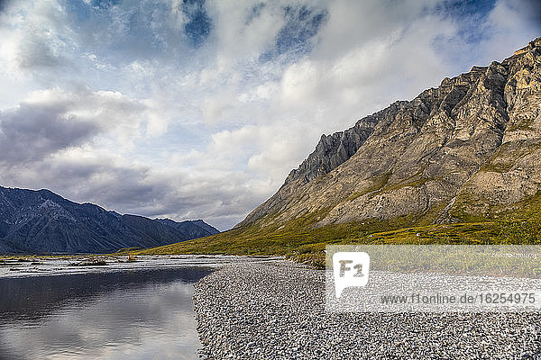 Rocky beach along the Marsh Fork river  with sunny mountains on the right side of the river  and dark stormy mountains on the left side of the river  while storm clouds dissipate  blue sky starting to peak through  Brooks Range  Arctic National Wildlife Refuge; Alaska  United States of America