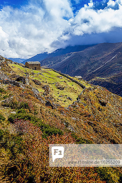 Verlassenes Sherpa-Haus  dessen Land von einer Felswand gesäumt ist  und Herbstfarben  die die Tundra erhellen  tauchen an diesem sonnigen Herbsttag aus den Wolken entlang des Trecks nach Phortse auf dem Gokyo-Trek auf  Sagarmantha-Nationalpark  Khumbu-Region  Nepal; Khumbu-Region  Nepal