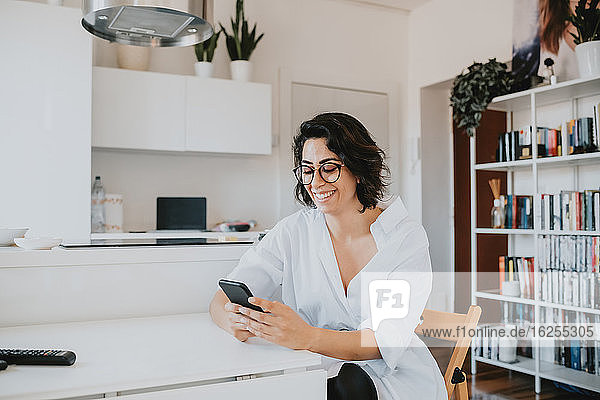 Woman with brown hair wearing glasses sitting at table in an apartment  using mobile phone.