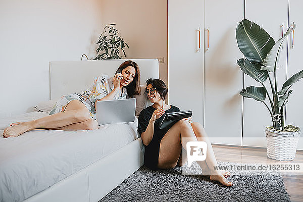 Two women with brown hair sitting on floor and lying on daybed  using laptop and mobile phone.