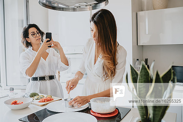 Two smiling women with brown hair standing in a kitchen  preparing food  taking picture with mobile phone.