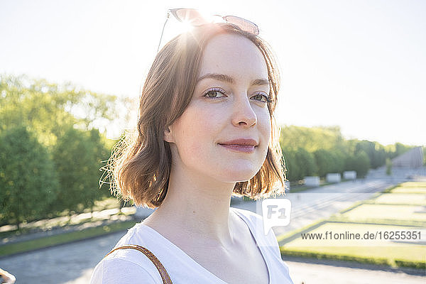 Porträt einer jungen Frau mit brünetten Haaren  die in einem Park steht und in die Kamera lächelt.