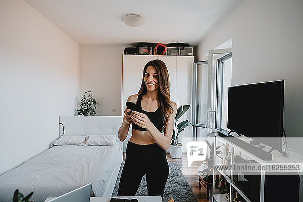 Frau mit braunen Haaren in Sportkleidung steht im Wohnzimmer und benutzt ein Mobiltelefon.