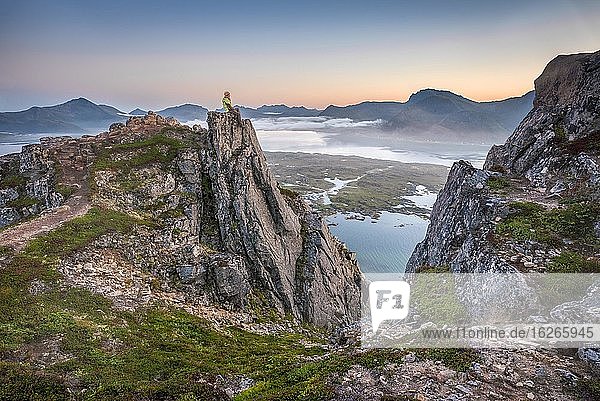 Frau sitzt auf Felsvorsprung Gipfel des Hoven bei Sonnenuntergang  hinten Meer und Fjord  Gimsøy  Lofoten  Nordland  Norwegen  Europa