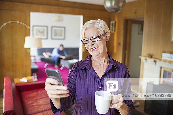 Glückliche ältere Frau benutzt Smartphone und trinkt Tee im Wohnzimmer