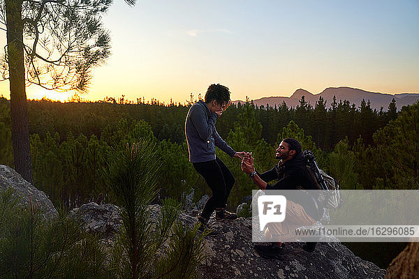 Junger Mann mit Verlobungsring macht der Frau bei Sonnenuntergang einen Heiratsantrag auf einem Felsen
