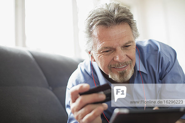 Älterer Mann mit Kopfhörer und Kreditkarte mit digitalem Tablett auf dem Sofa