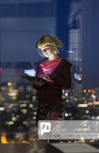 Junge Geschäftsfrau mit digitalem Tablet arbeitet spät am Bürofenster