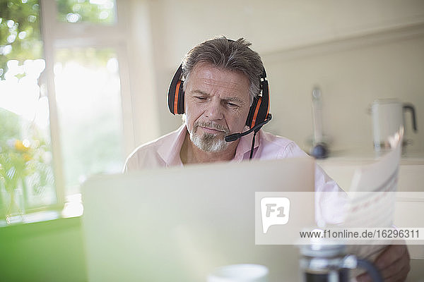 Älterer Mann mit Kopfhörern und Zeitung arbeitet am Laptop in der Küche