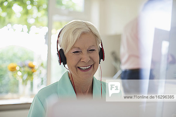 Glückliche ältere Frau mit Kopfhörern und Laptop in der Küche