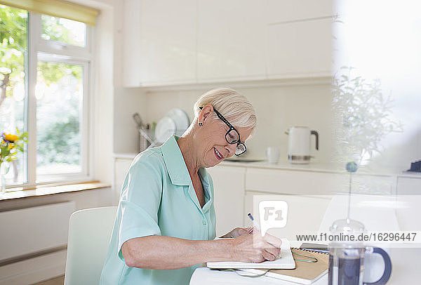 Ältere Frau schreibt am morgendlichen Küchentisch in einem Journal