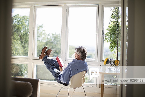 Sorgloser älterer Mann liest mit den Füßen am sonnigen Fenster