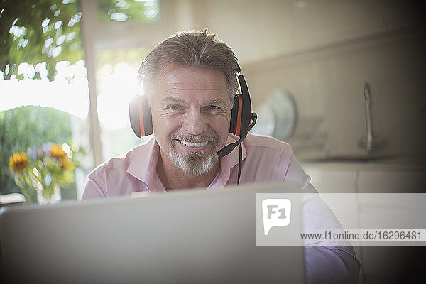 Porträt eines lächelnden älteren Mannes mit Kopfhörern bei der Arbeit am Laptop