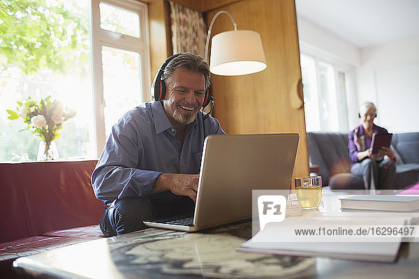 Glücklicher älterer Mann mit Kopfhörern und Laptop im Wohnzimmer