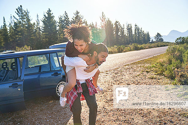 Verspieltes junges Paar beim Huckepackfahren im Auto am sonnigen  abgelegenen Straßenrand