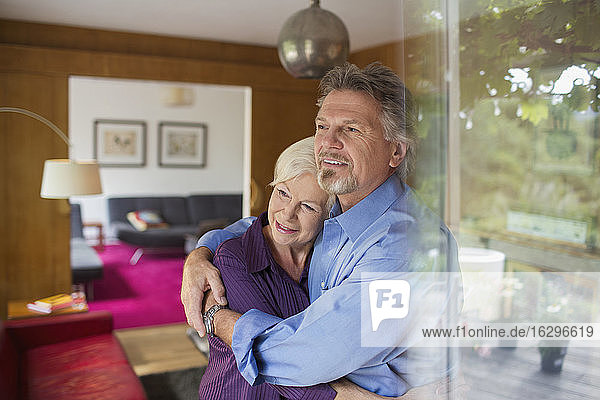 Glückliches älteres Paar umarmt sich am sonnigen Wohnzimmerfenster