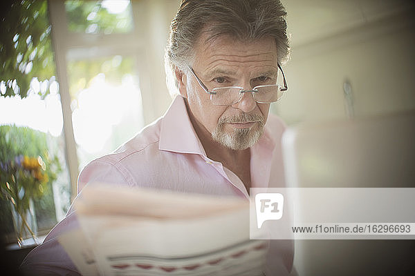 Älterer Mann liest Zeitung am Laptop in sonniger Morgenküche