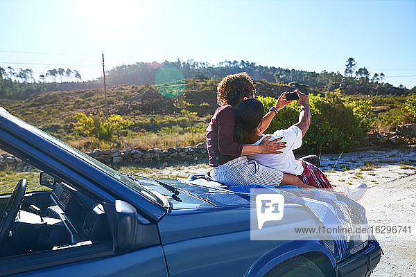 Sorgenfreies junges Paar  das sich am sonnigen Straßenrand auf der Motorhaube eines Autos selbstständig macht