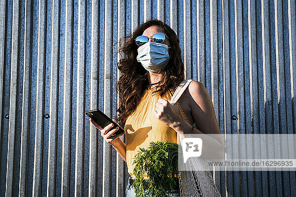 Frau mit Gesichtsmaske  während sie mit Mobiltelefon und wiederverwendbarer Netztasche an einer Metallwand steht