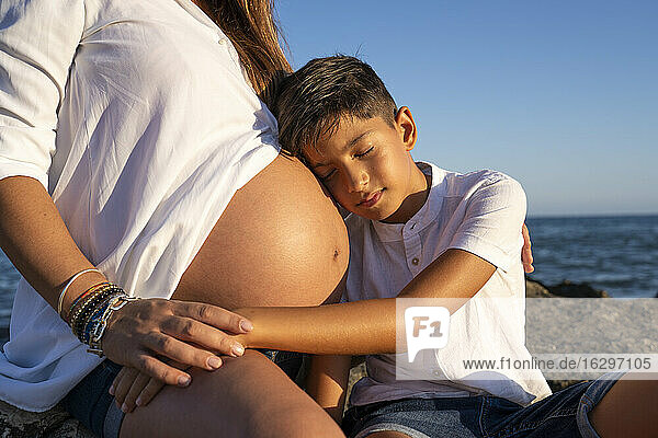 Sohn hält den Kopf auf dem Bauch der schwangeren Mutter  während er am Strand sitzt