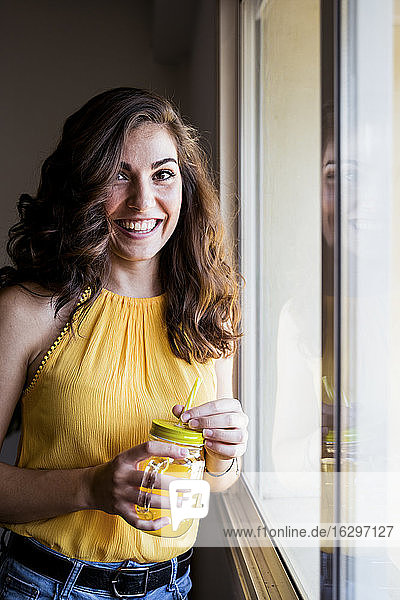 Schöne junge Frau hält Einmachglas mit Saft und lächelt am Fenster zu Hause
