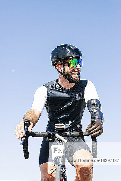 Male amputee cyclist laughing while riding bicycle against clear sky