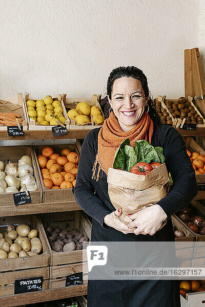 Gut gelaunte Verkäuferin  die eine Tüte mit Gemüse in der Hand hält  die vor Kisten im Laden steht