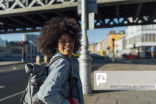 Lächelnde junge Frau mit Afro-Haar  die wegschaut  während sie auf dem Bürgersteig an einem sonnigen Tag steht