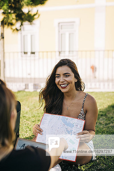 Fröhliche Frau zeigt einem Freund eine Karte  während sie auf einer Wiese sitzt