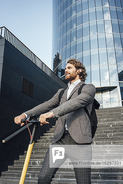 Gutaussehender männlicher Fachmann  der mit einem Elektroroller vor einem Bürogebäude steht und wegschaut