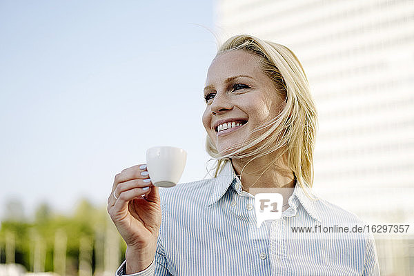 Smiling blond female professional holding coffee cup while looking away against sky