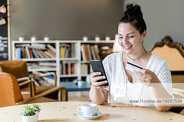 Smiling woman with coffee on table doing online shopping over mobile phone in cafe