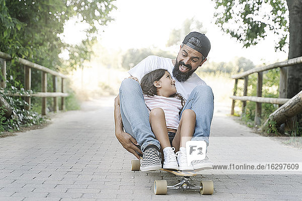 Fröhlicher Vater und Tochter beim Skateboardfahren auf dem Fußweg im Park
